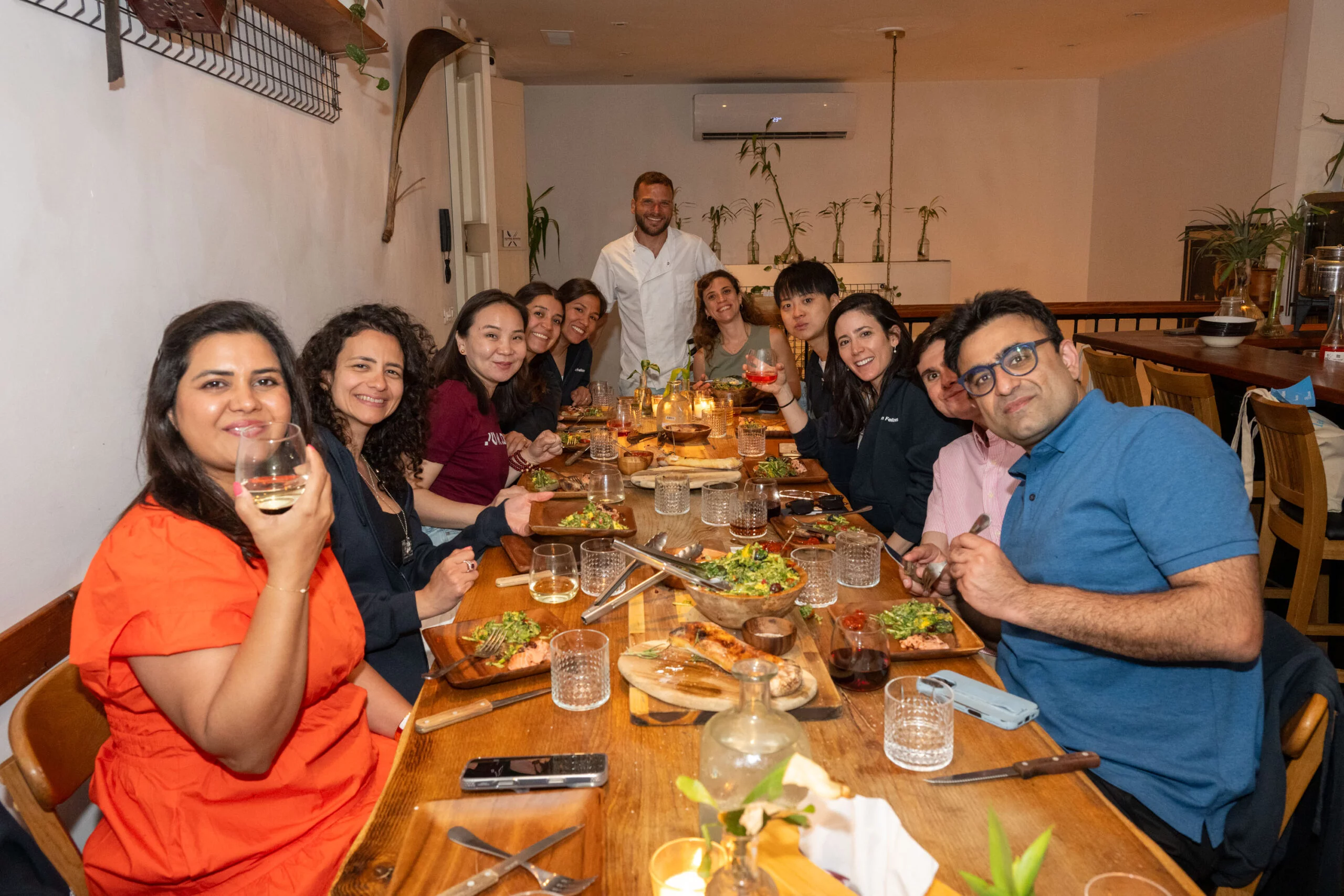 ChatGPT said: קבוצה של סועדים סביב שולחן עץ ארוך במסעדת "מעשנת השוק", נהנים מארוחת שף מעושנת עם יין ומנות צבעוניות.